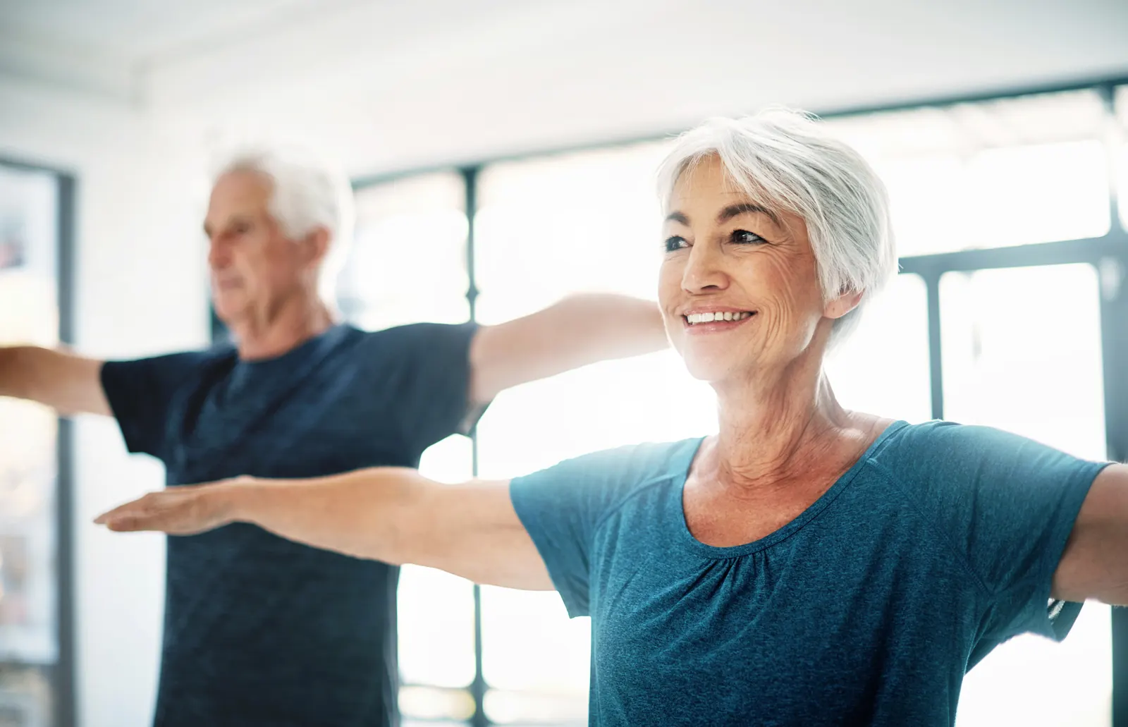 Un couple de seniors souriants pratique des exercices d'étirement, bras tendus, dans une salle lumineuse.