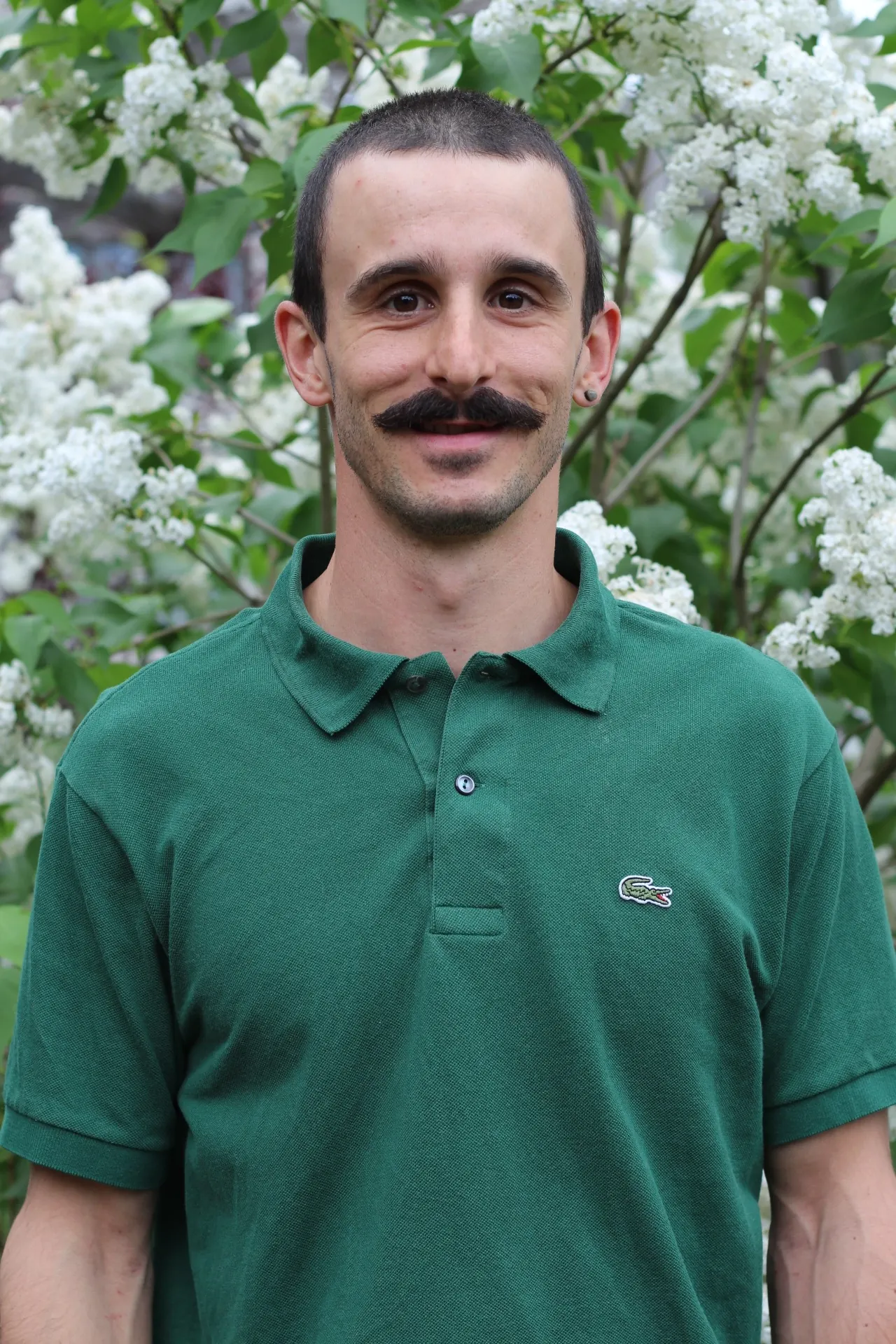Portrait d'Enzo His, ostéopathe, souriant, en polo vert, devant un lilas blanc en fleurs.